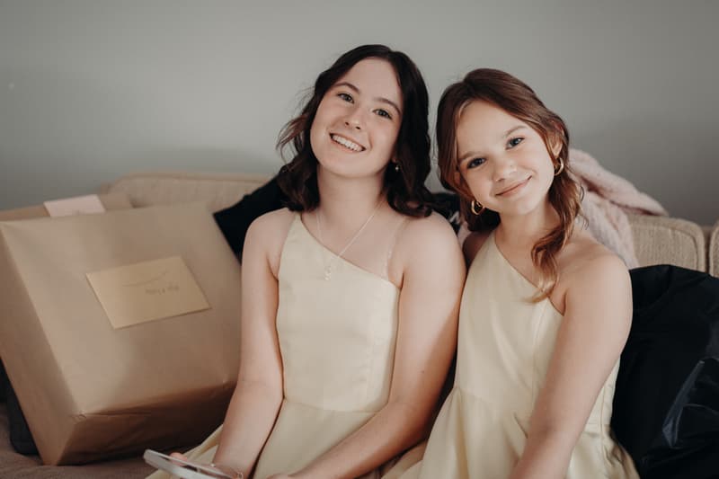 Two young bridesmaids sit on a couch smiling, wearing matching cream-colored dresses.