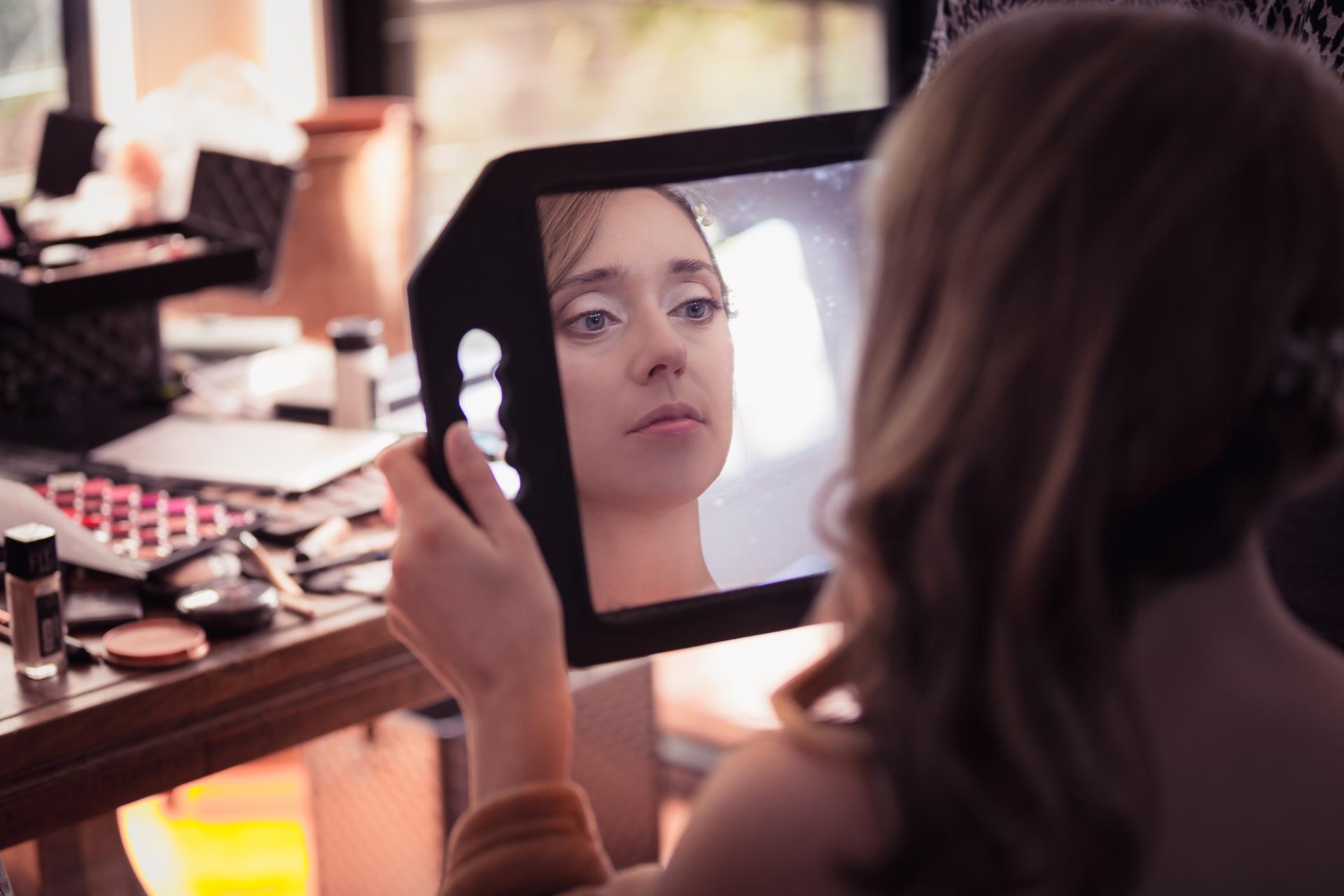 The bride Francesca holds a mirror reflecting her face while seated at a table with makeup products during bridal preparation.