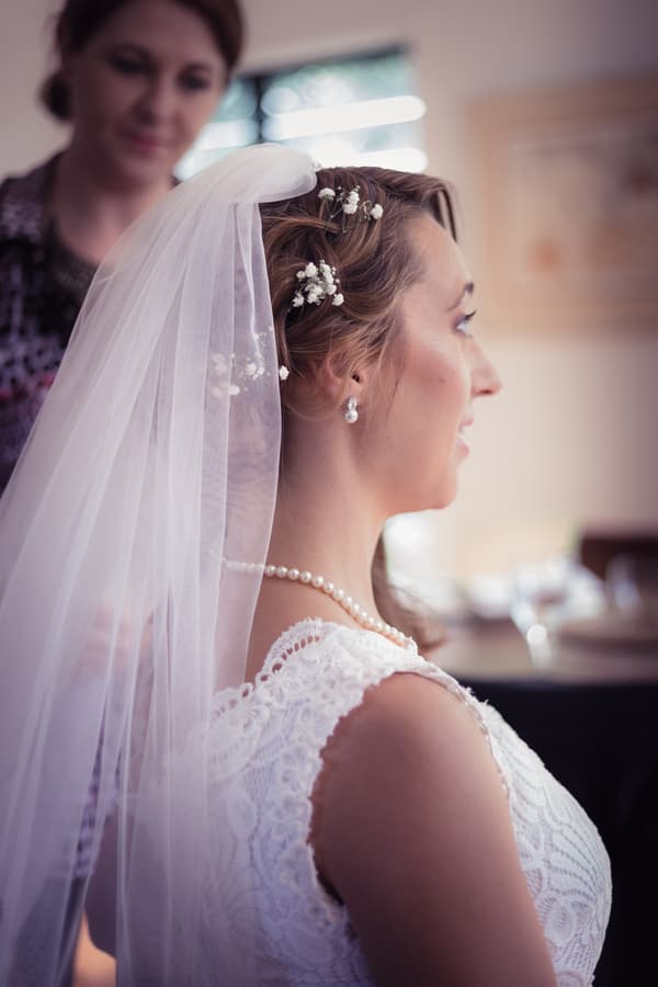 The bride Francesca is seated wearing a lace wedding dress, pearl necklace, and veil adorned with small flowers, while another woman stands behind her indoors.