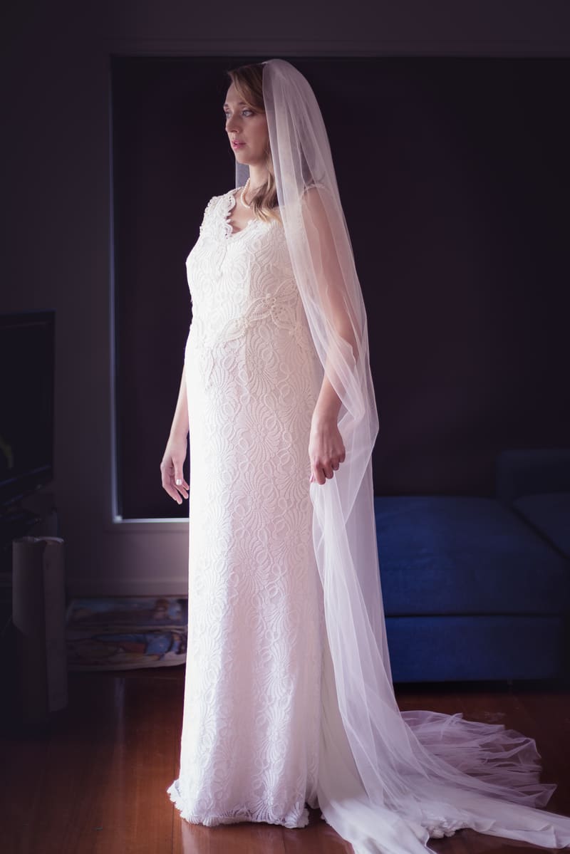 The bride Francesca stands indoors wearing a lace wedding gown and long veil, looking to the side. Engagment party by She Said Yes.