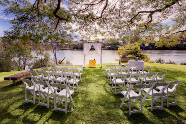 Empty outdoor wedding ceremony setup at TOOWONG ROWING CLUB by the Brisbane River's edge, featuring white folding chairs arranged in rows facing a small altar with a white draped arch and a yellow tablecloth.