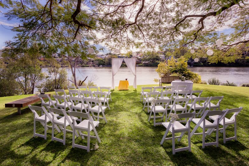 Empty outdoor wedding ceremony setup at TOOWONG ROWING CLUB by the Brisbane River's edge, featuring white folding chairs arranged in rows facing a small altar with a white draped arch and a yellow tablecloth.