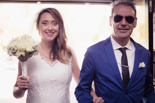 The bride Francesca, holding a bouquet of white flowers, stands next to an older man in a blue suit and sunglasses, likely her father, at the entrance of Toowong Rowing Club.
