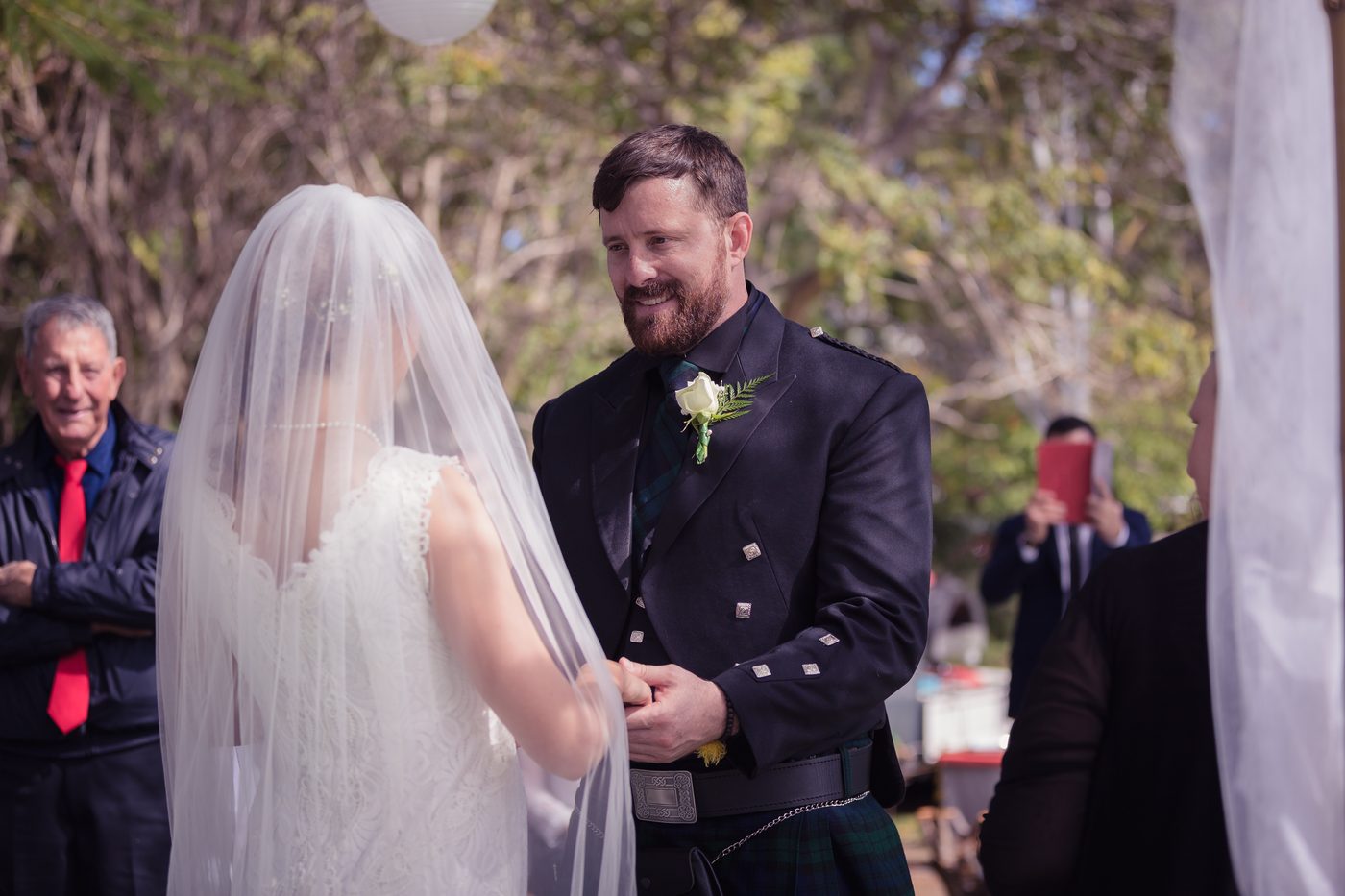 Francesca the bride and Ben the groom hold hands during their wedding ceremony, with guests visible in the background. Destination wedding photographer Australia by She Said Yes.