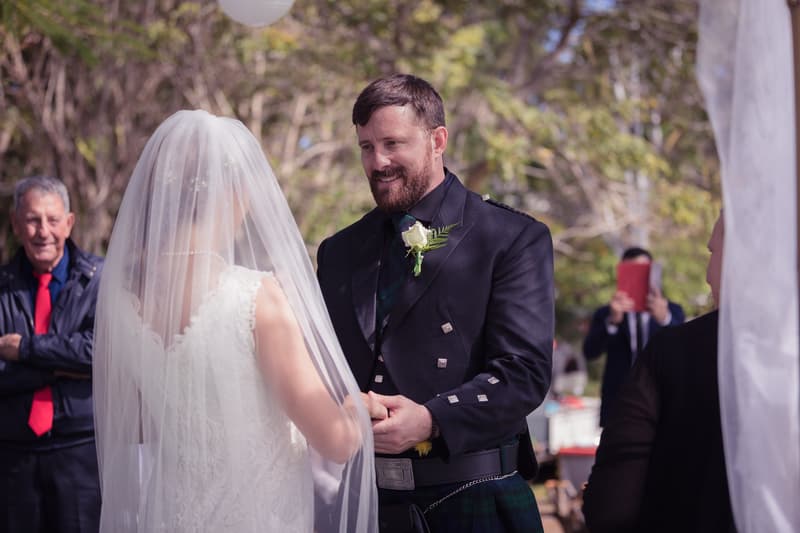 Francesca the bride and Ben the groom hold hands during their wedding ceremony at TOOWONG ROWING CLUB, with guests visible in the background.