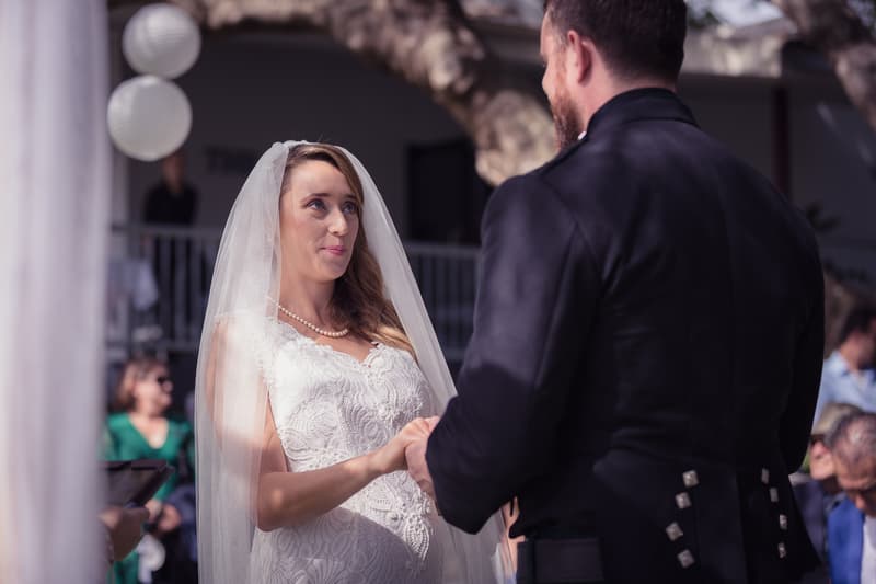 The bride Francesca and groom Ben hold hands facing each other during their wedding ceremony at Toowong Rowing Club by the Brisbane River's edge, with guests seated in the background.