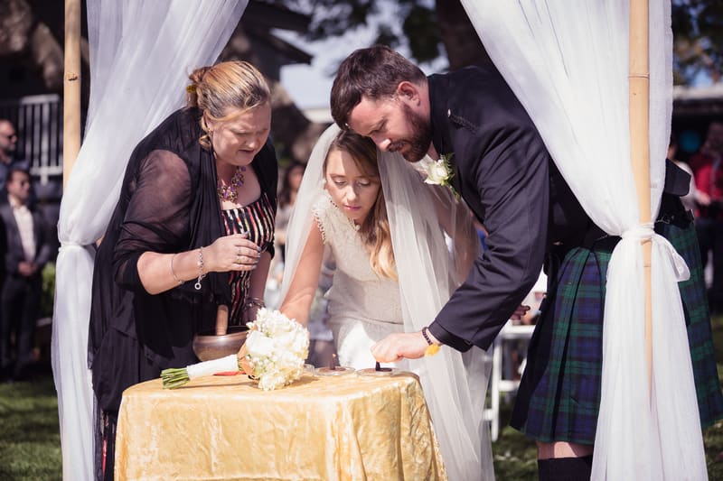 The bride Francesca and groom Ben, accompanied by an older woman, participate in a ceremonial ritual at a table covered with a gold cloth. Full day wedding photography by She Said Yes.