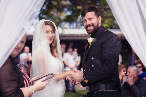 Bride Francesca and groom Ben hold hands during their wedding ceremony, with an officiant standing nearby and guests seated in the background. Engagment party by She Said Yes.