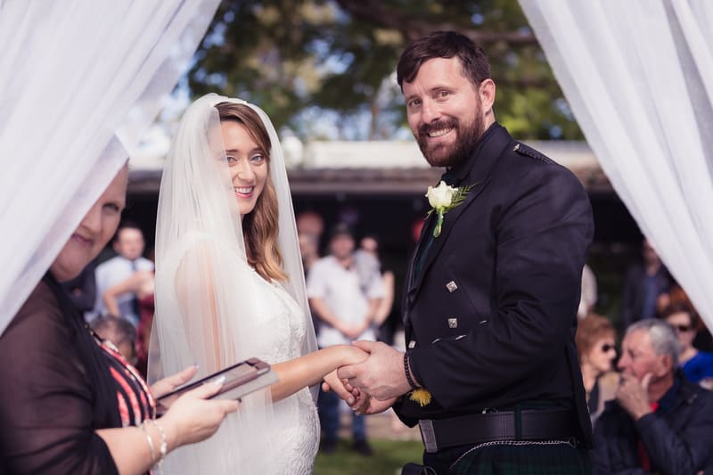 Bride Francesca and groom Ben hold hands during their wedding ceremony, with an officiant standing nearby and guests seated in the background. Destination wedding video packages by She Said Yes.