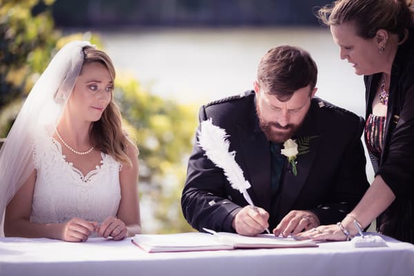 The bride Francesca watches as the groom Ben signs the wedding register with a white feather pen, assisted by a woman standing beside him. Brisbane wedding video by She Said Yes.
