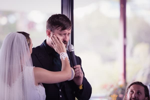 Bride Francesca gently touches the face of groom Ben, who is holding a microphone and appears emotional, during the reception at TOOWONG ROWING CLUB — The Malouf Room.