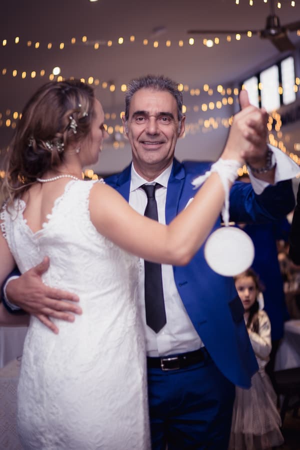 The bride Francesca dances with an older man, likely her father, at the reception in The Malouf Room at Toowong Rowing Club. Francesca wears a white lace wedding dress and a pearl necklace, while the man wears a blue suit with a black tie. String lights decorate the ceiling in the background.