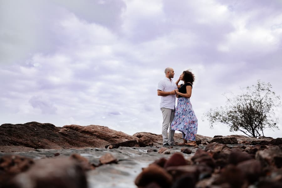 The couple stands on rocky terrain at Shorncliffe beachfront, Brisbane, under a cloudy sky, holding hands and gazing at each other.