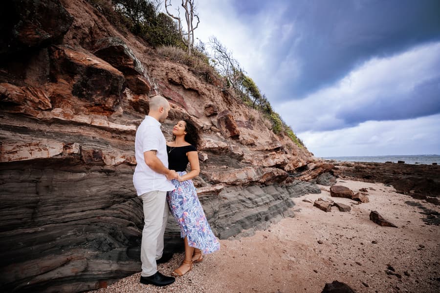 The couple embraces in front of a layered rock formation at Shorncliffe beachfront, with a dramatic cloudy sky above.