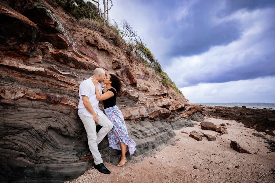 The couple shares a kiss in front of a textured rock wall at Shorncliffe beachfront, with the ocean visible in the background.