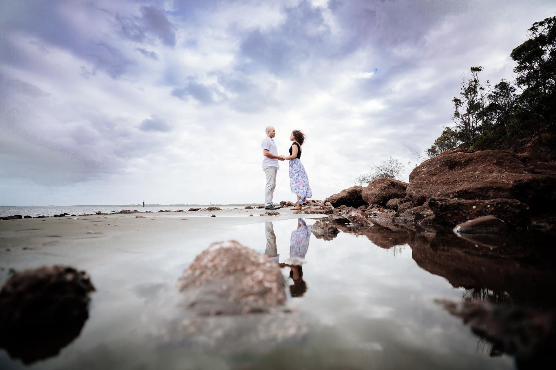 The couple stands on wet sand at Shorncliffe beachfront, reflected in a shallow pool of water, with a cloudy sky above.
