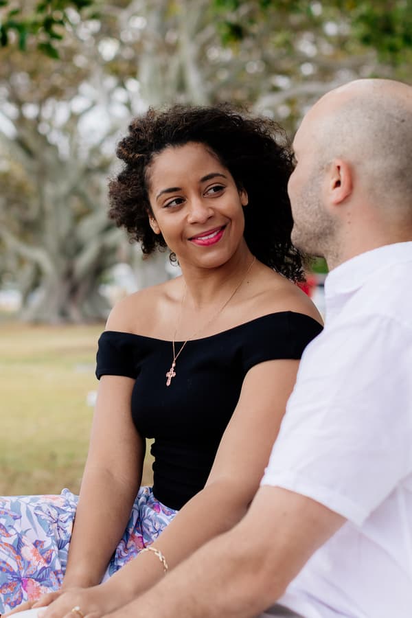 The couple sits on grass at Shorncliffe beachfront, smiling at each other with trees in the background.