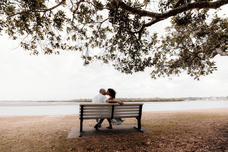 The couple sits on a bench under a large tree at Shorncliffe beachfront, looking out over the water.