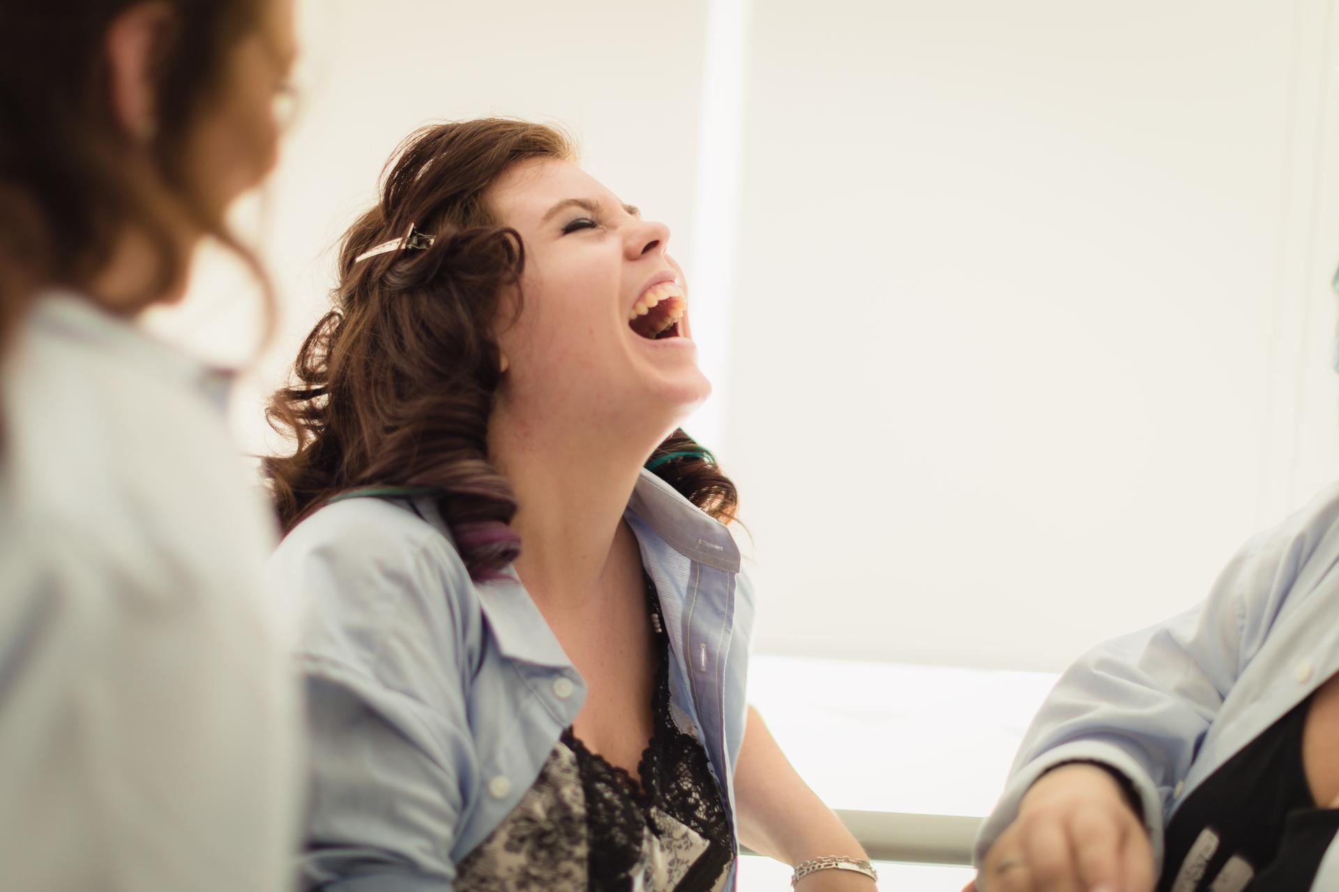 The bride is laughing joyfully while wearing a partially open shirt over a lace top, with two other people partially visible nearby.