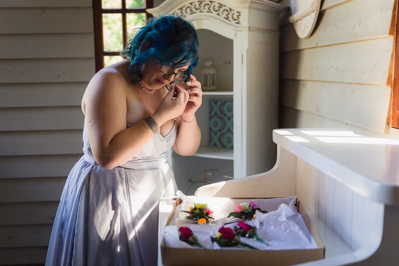 The bride Holly adjusts her earring while standing next to a white wooden cabinet with a box of boutonnieres and floral accessories at Kwila Lodge.