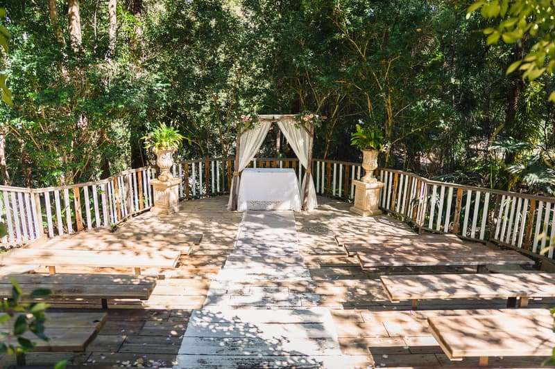 Empty outdoor ceremony stage at Kwila Lodge with wooden benches, a white aisle runner, and a small altar decorated with draped fabric and floral arrangements.