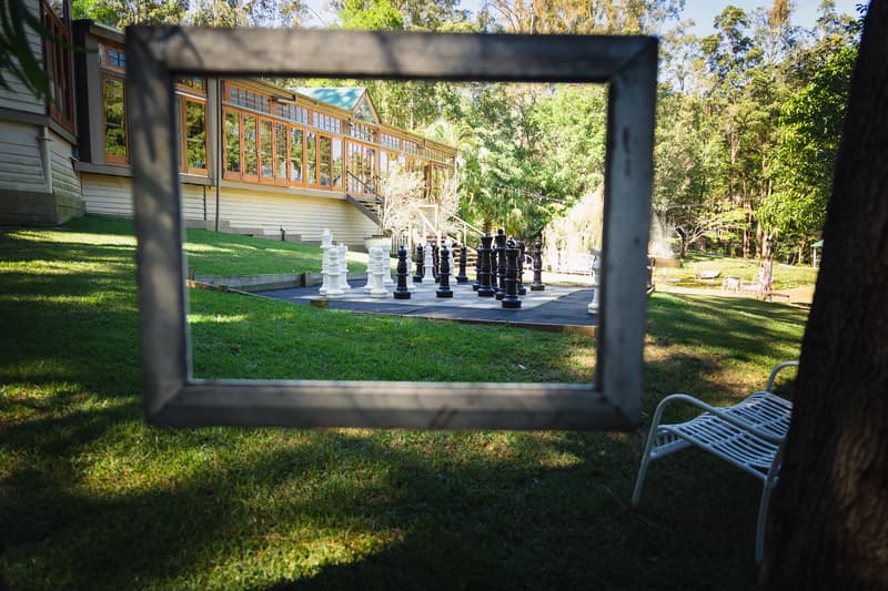 Outdoor area at Kwila Lodge showing a large chessboard with oversized chess pieces framed by a hanging rectangular frame, with a white metal chair and lodge building in the background.