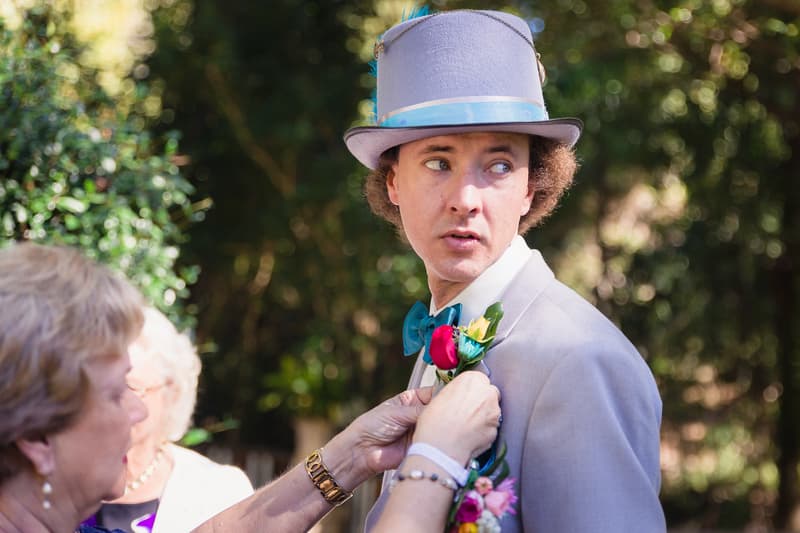 The groom Paul wearing a light grey suit, blue bow tie, and grey top hat with a blue ribbon has a boutonniere pinned on his lapel by an older woman, likely the mother of the groom, at Kwila Lodge.