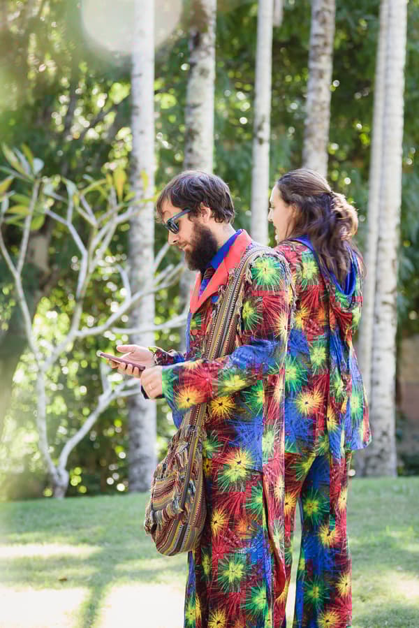 The bride and groom stand outdoors at Kwila Lodge wearing matching colorful, patterned outfits with starburst designs; the groom is looking at a phone while the bride faces away.