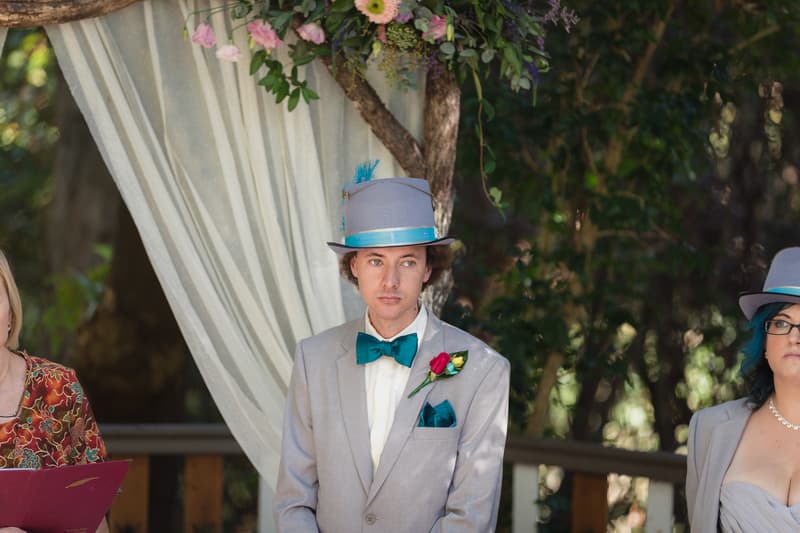 The groom Paul stands at the ceremony stage at Kwila Lodge wearing a light grey suit, teal bow tie, and a grey top hat with a teal band and feather, with floral decorations and draped fabric behind him.