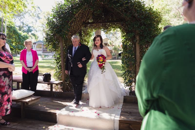 The bride Holly, holding a bouquet, walks arm-in-arm with an older man, likely her father, under a leafy arch at Kwila Lodge during the ceremony. Guests stand nearby watching.