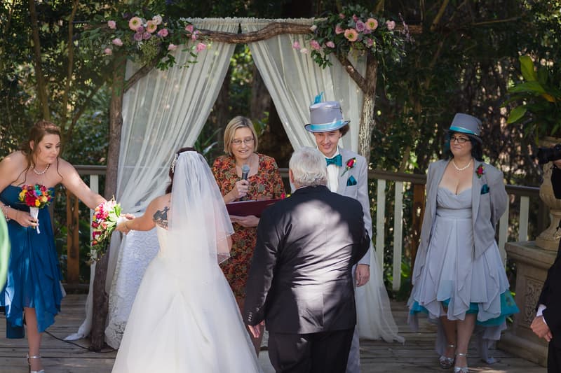 Bride Holly is being given away by an older man, likely her father, at the ceremony stage at Kwila Lodge. The officiant stands under a floral arch with white drapes, reading from a book. Groom Paul, wearing a grey suit and top hat with a teal bow tie, stands smiling nearby. A bridesmaid in a blue dress holds a bouquet and reaches out to the bride. Another woman in a grey dress and matching hat stands to the side.