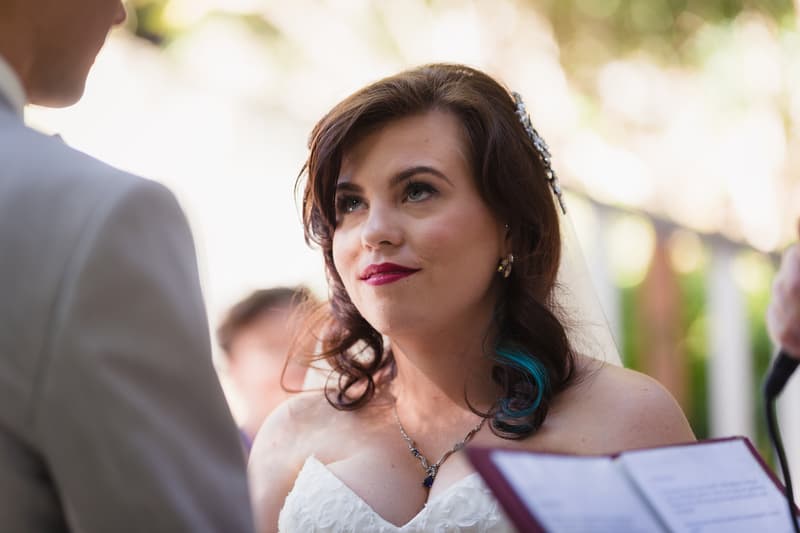 The bride Holly looks up at the groom Paul during their wedding ceremony at Kwila Lodge, with an officiant holding a book visible on the right.