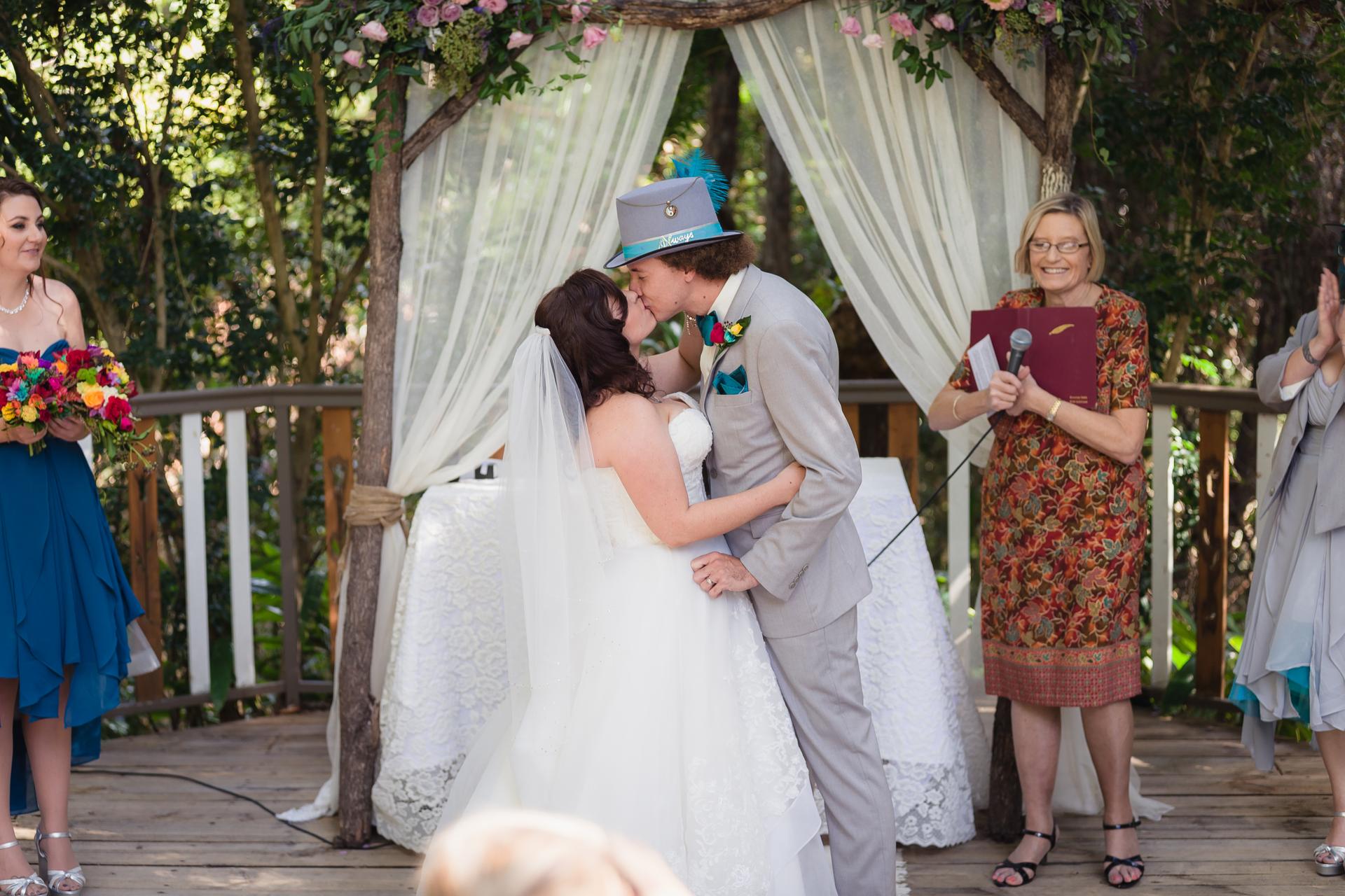 Bride Holly and groom Paul share a kiss at the altar during their wedding ceremony at Kwila Lodge, with the officiant holding a microphone and guests including a bridesmaid holding a bouquet nearby.