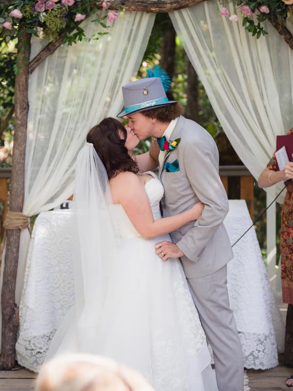Bride Holly and groom Paul share a kiss at the altar during their wedding ceremony at Kwila Lodge, with the officiant holding a microphone and guests including a bridesmaid holding a bouquet nearby.