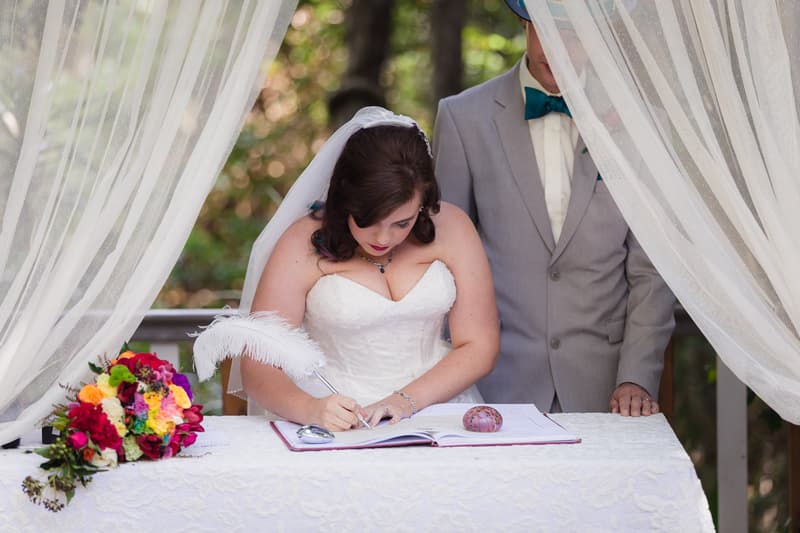 The bride Holly signs the wedding register at Kwila Lodge during the ceremony, while the groom Paul stands beside her. A colorful bridal bouquet rests on the table covered with a white lace cloth.