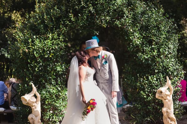 Bride Holly and groom Paul kiss under a leafy arch at Kwila Lodge during their wedding ceremony, with guests seated in the background and decorative statues on either side of the arch.