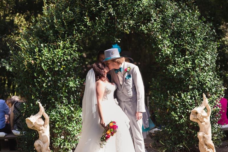 Bride Holly and groom Paul kiss under a leafy arch at Kwila Lodge during their wedding ceremony, with guests seated in the background and decorative statues on either side of the arch.