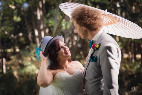 The bride wearing a white strapless wedding gown and holding a grey hat with a blue feather looks up at the groom, who is dressed in a light grey suit with a blue bow tie and colorful boutonniere, holding a white parasol. Candid wedding photographer by She Said Yes.