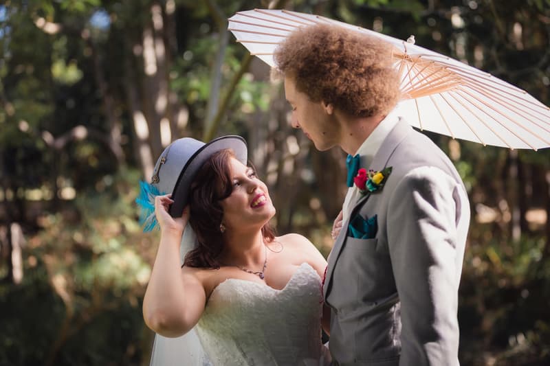 The bride wearing a white strapless wedding gown and holding a grey hat with a blue feather looks up at the groom, who is dressed in a light grey suit with a blue bow tie and colorful boutonniere, holding a white parasol at Kwila Lodge outdoors.