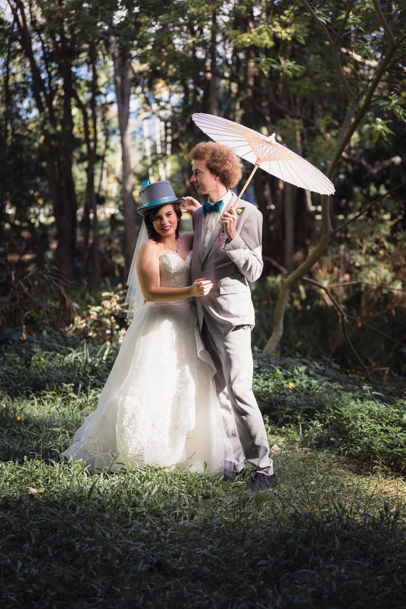 The bride Holly and groom Paul pose outdoors in a wooded area. Holly wears a white wedding gown and a blue top hat, while Paul wears a light grey suit with a teal bow tie and holds a white parasol. Wedding fair season Brisbane by She Said Yes.