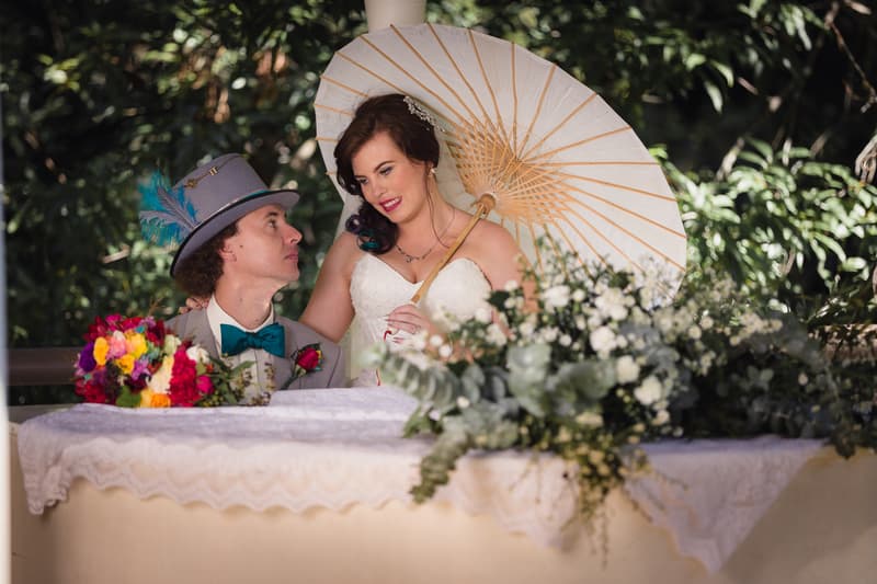 Bride Holly holds a white parasol and looks at groom Paul, who wears a grey suit with a teal bow tie and feathered hat, seated behind a lace-covered table with floral arrangements at Kwila Lodge.