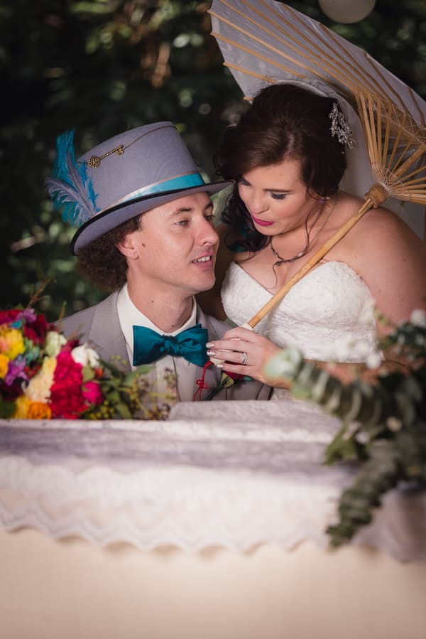 Bride Holly and groom Paul pose closely together at Kwila Lodge, with Holly holding a parasol and Paul wearing a grey hat with blue feathers and a teal bow tie.