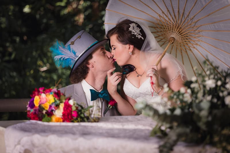 Bride Holly and groom Paul share a kiss at Kwila Lodge, with Holly holding a parasol and Paul wearing a grey suit and feathered hat. A colorful bouquet is visible in the foreground.