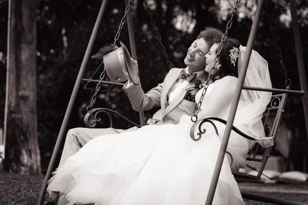 Bride Holly and groom Paul sit together on a swing outdoors, with Paul holding his hat in the air and both smiling. Engagement parties by She Said Yes.