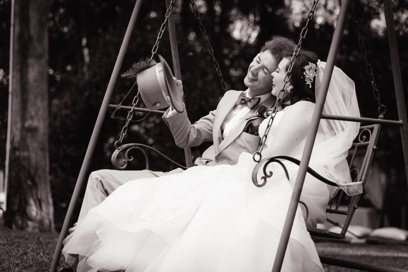 Bride Holly and groom Paul sit together on a swing outdoors, with Paul holding his hat in the air and both smiling.