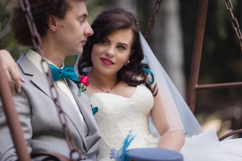 Bride Holly and groom Paul sit closely together on a swing at Kwila Lodge, with Holly looking at Paul while he looks downward. Holly wears a white wedding dress and veil, and Paul wears a light grey suit with a teal bow tie and a red boutonniere.