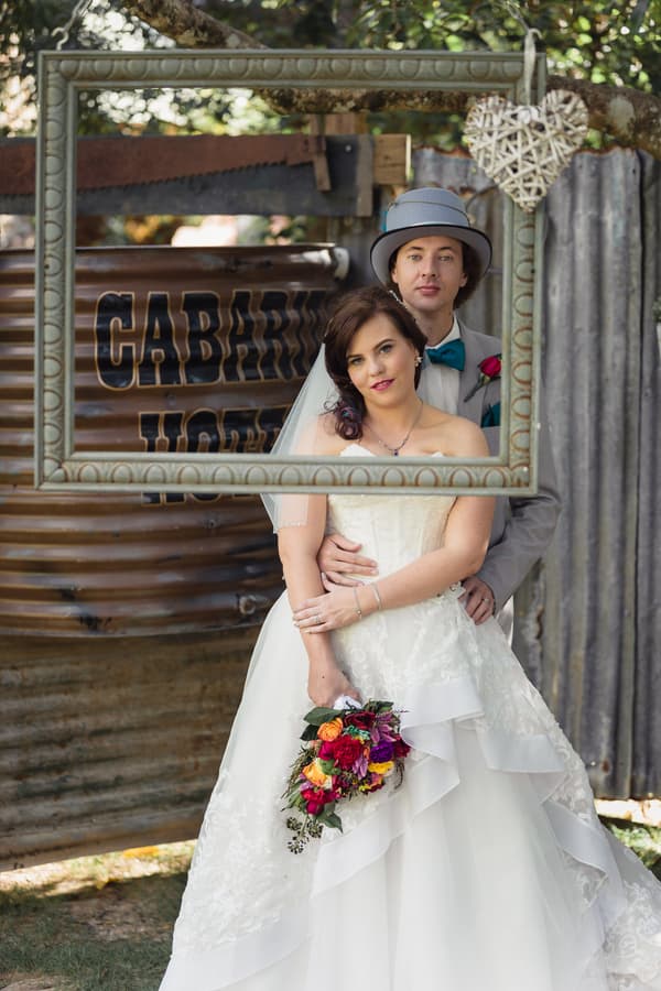 Bride Holly and groom Paul pose together framed by a hanging picture frame at Kwila Lodge, with a rustic corrugated metal backdrop featuring a sign partially reading 'CABAR... LODGE'. Holly holds a colorful bouquet of flowers.