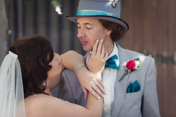Bride Holly in a veil and groom Paul in a grey suit with teal bow tie and hat pose closely together. Wedding live streaming by She Said Yes.