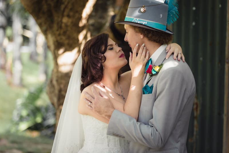 Bride Holly and groom Paul embrace outdoors near a tree at Kwila Lodge, with Holly wearing a white wedding dress and veil, and Paul in a light grey suit with a feathered hat.