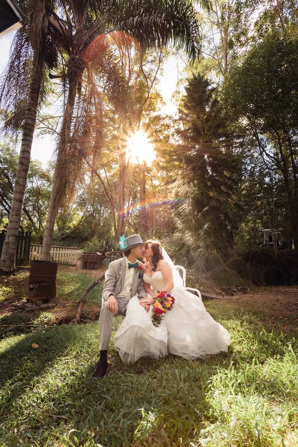 Bride Holly and groom Paul sit on a white chair outdoors at Kwila Lodge, sharing a kiss with the sun shining through tall trees behind them. Holly holds a colorful bouquet while wearing a white wedding gown, and Paul wears a light gray suit with a blue bow tie and a gray hat with a blue feather.